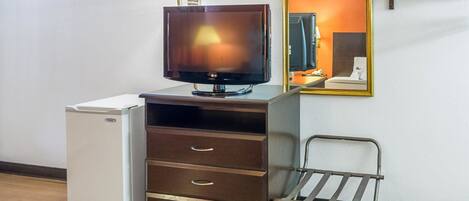 Habitación compacta con televisión de pantalla plana, un refrigerador pequeño, un mueble de madera con cajones, una silla de metal y un espejo en la pared.