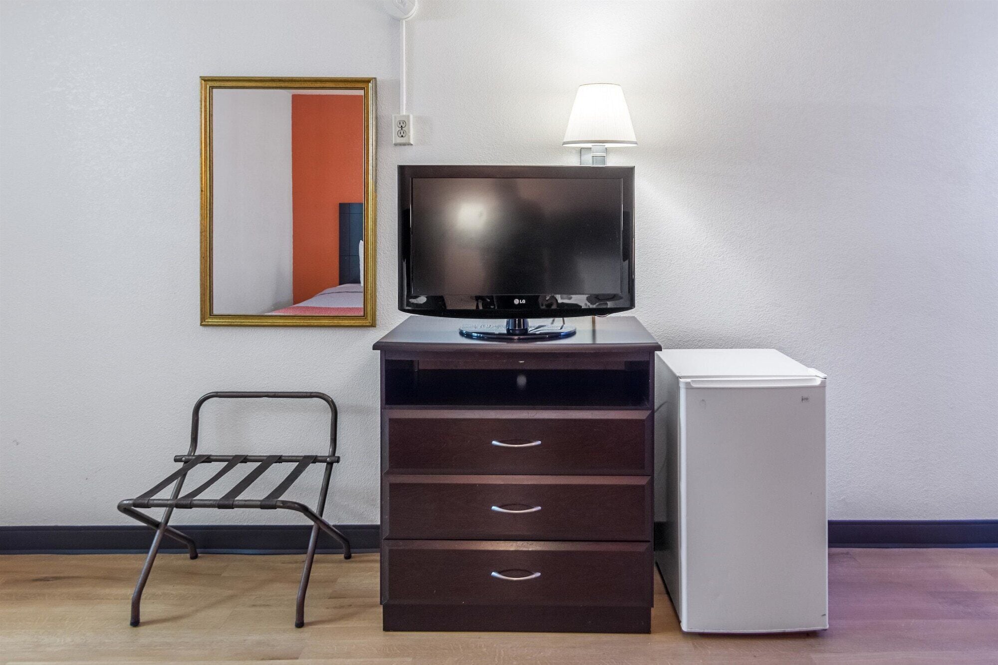 A room with a flat-screen TV on a wooden cabinet, a small white refrigerator, a metal chair, and a mirror on the wall.
