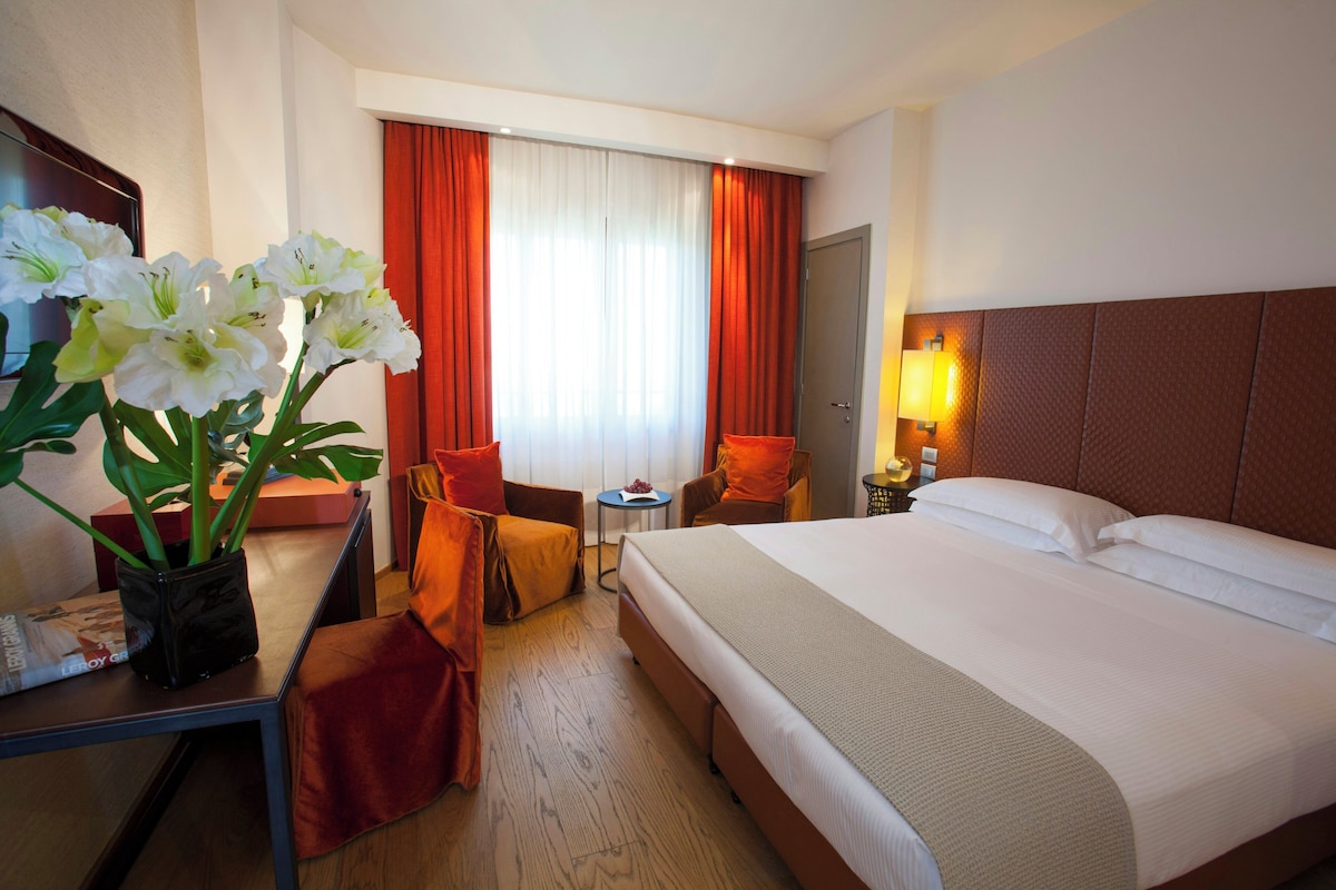 A hotel room with a bed, a desk with a chair, a small table, and a potted plant.