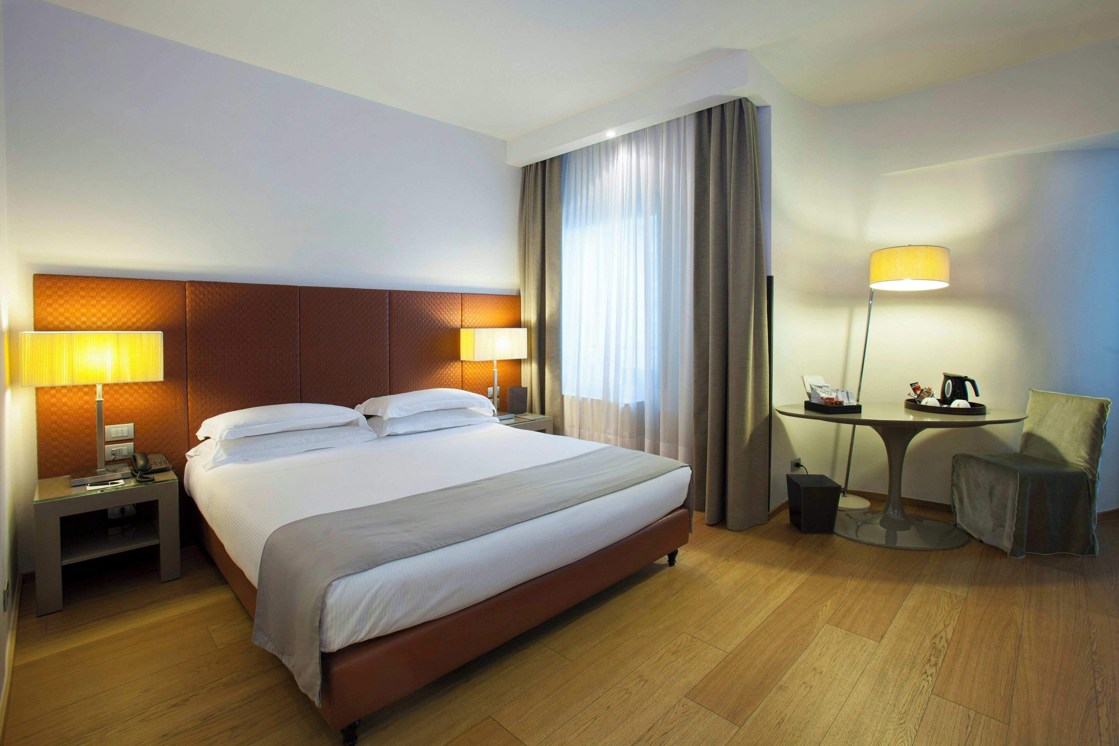 A hotel room with a large bed, two bedside lamps, a small table with a lamp, a chair, and a desk with a telephone.