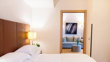 Habitación de hotel moderna con una cama grande, cabecera de madera, mesita de noche con lámpara y vista al área de estar por una puerta abierta.