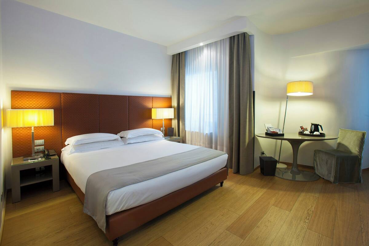 A hotel room with a large bed, two bedside lamps, a small table with a lamp, a chair, and a desk with a telephone.