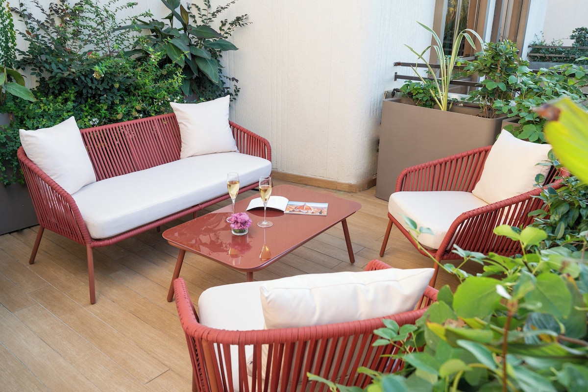 A red wicker sofa with white cushions, a matching red armchair, and a small table with a glass of wine and a book.