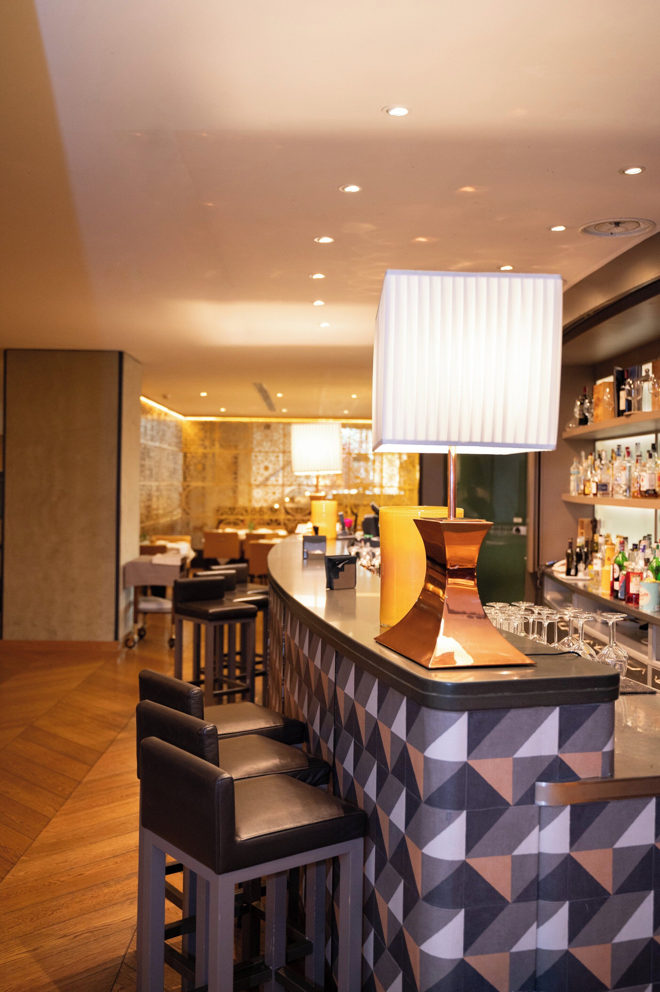 Un espace bar moderne avec un comptoir au design géométrique, des tabourets de bar rembourrés et une grande suspension blanche.