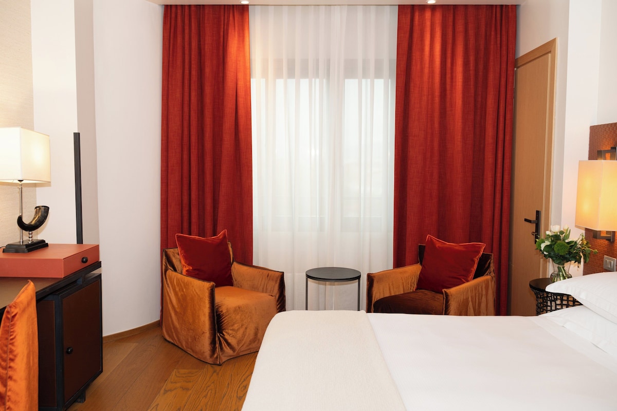 A hotel room with a bed, two armchairs, a small table, a desk with a lamp, and a door.