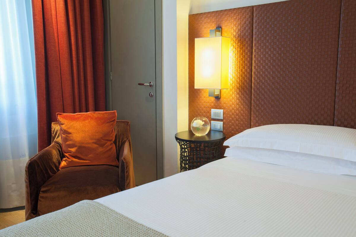 A hotel room with a bed, a chair, a nightstand with a lamp, and a wall-mounted light fixture.