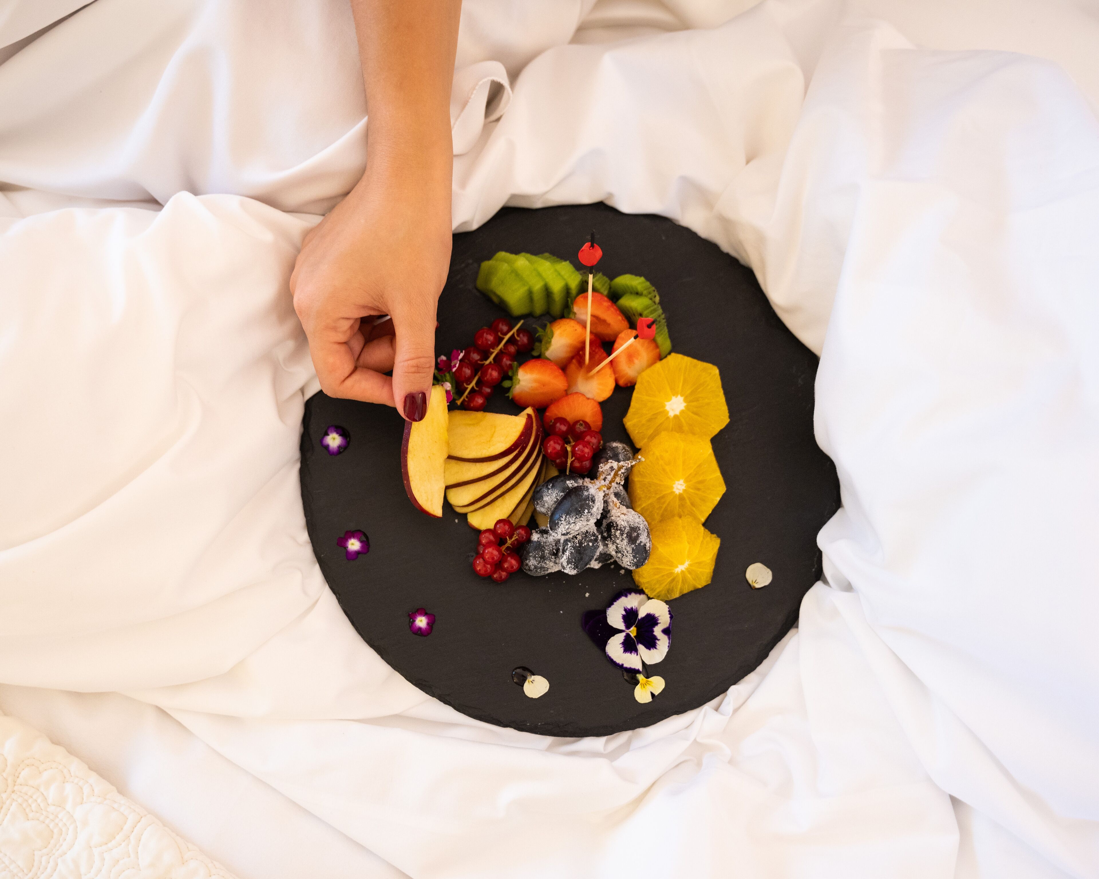 Una mano che tiene una fetta di mela su un piatto nero con diversi frutti e fiori.