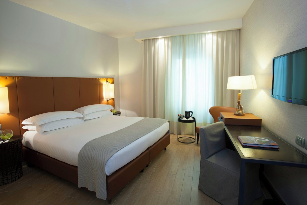 A hotel room with a large bed, a desk with a chair, a television, and a window with curtains.