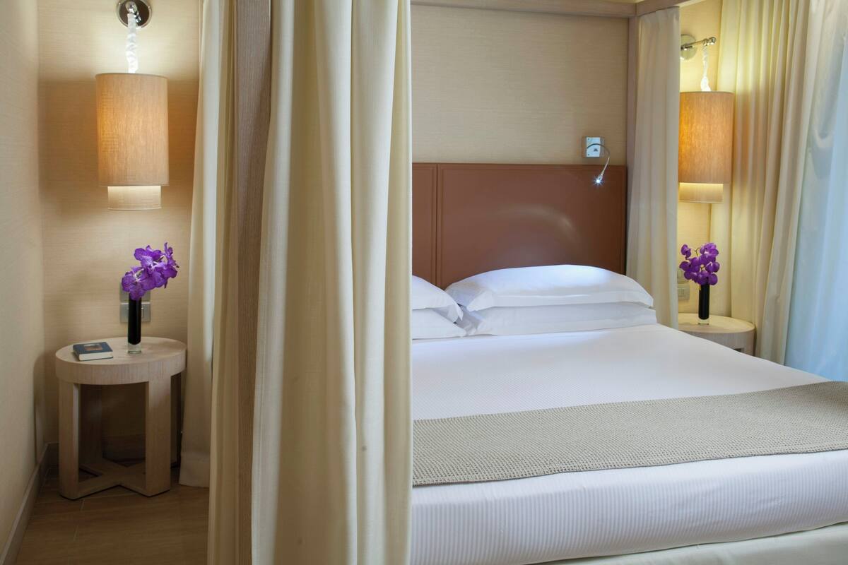 A hotel room with a bed, two bedside lamps, a round table with a book and a vase of flowers, and a wall-mounted lamp.