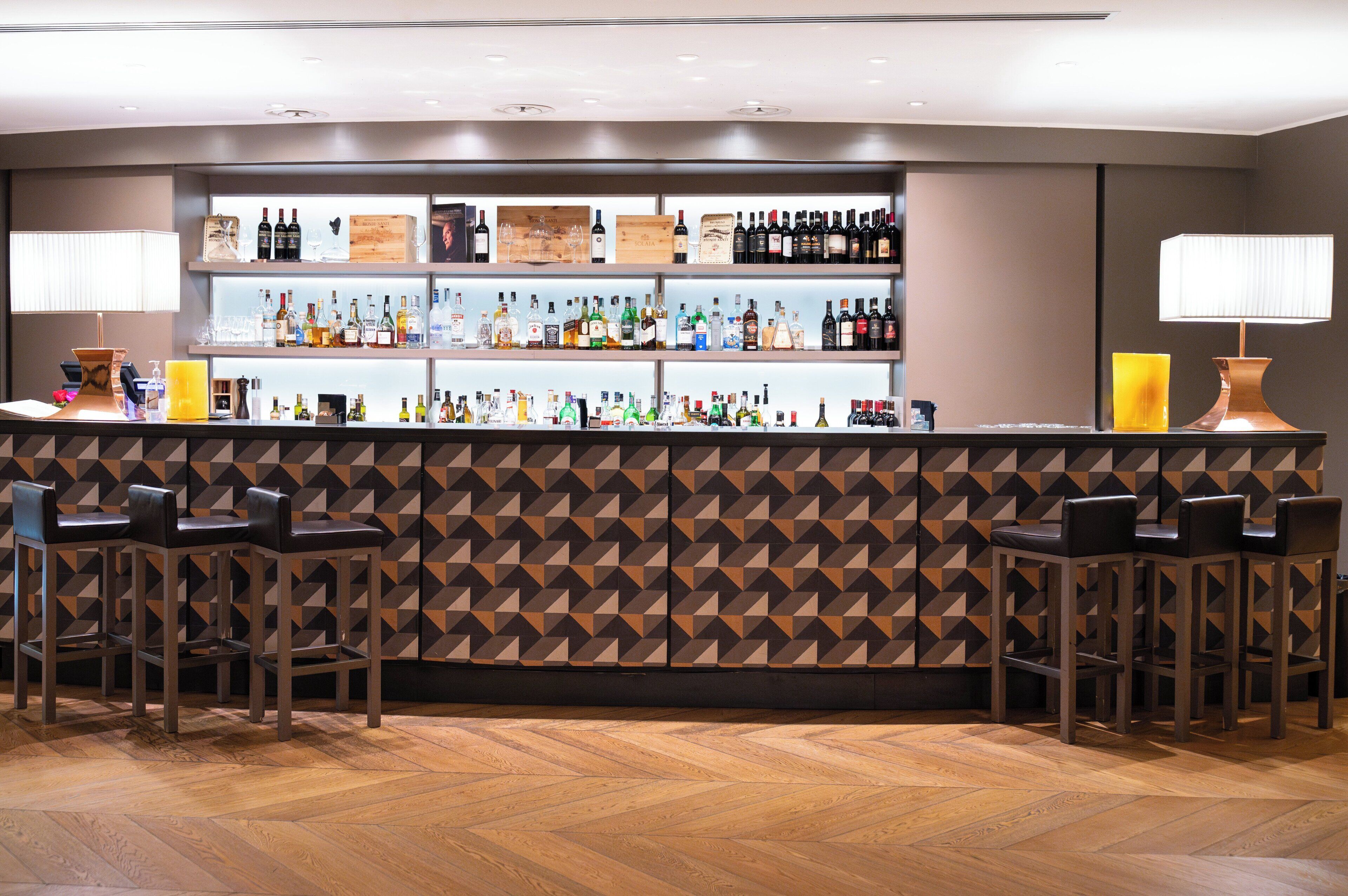 A bar area with a geometric patterned wall, multiple shelves of liquor bottles, and bar stools.