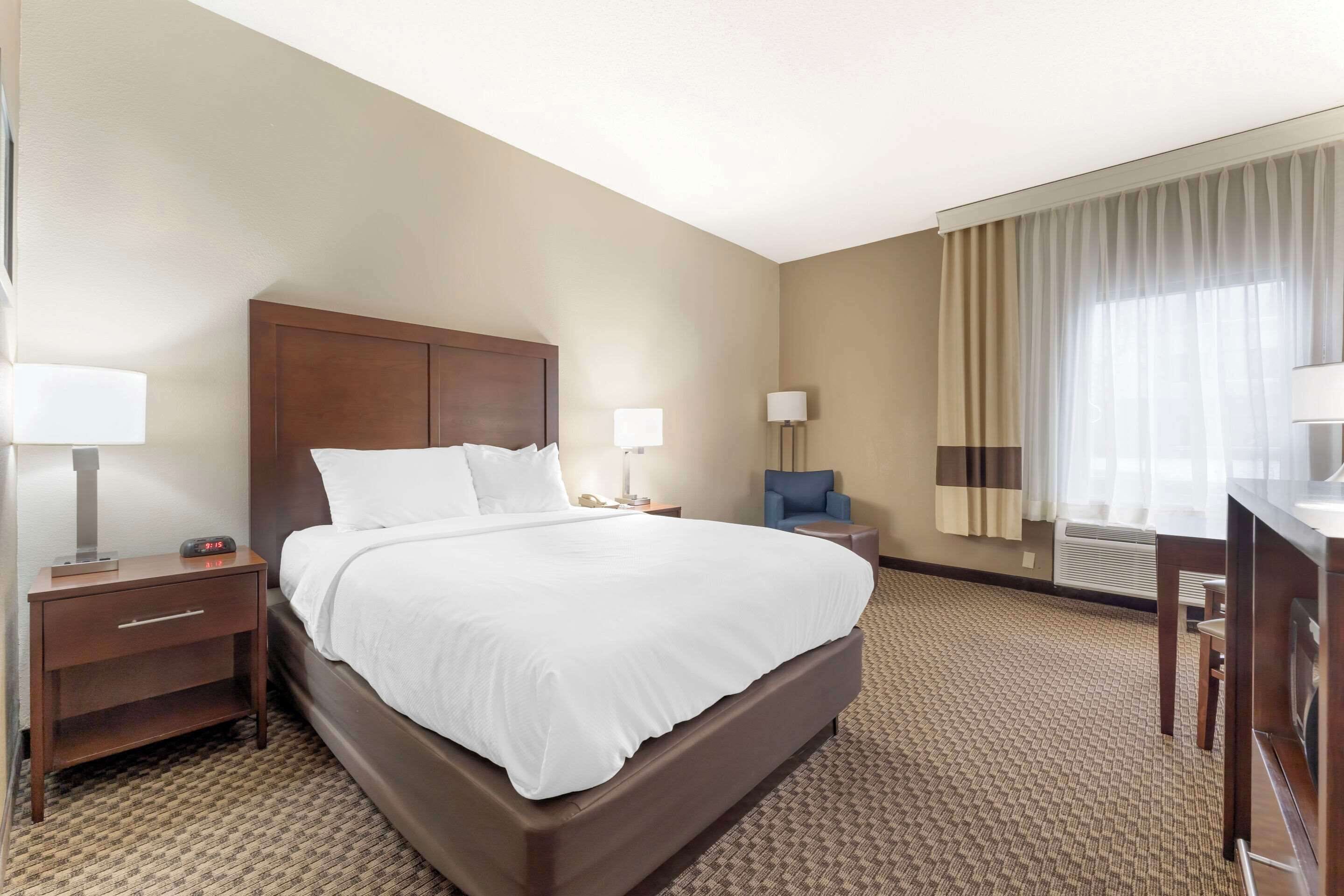 Ein Hotelzimmer mit einem großen Bett, einem Nachttisch mit Lampe, einem Schreibtisch mit Stuhl und einem Fenster mit Vorhängen.