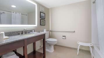 Casa de banho com lavatório, sanita e chuveiro. Há um banquinho branco e um corrimão.