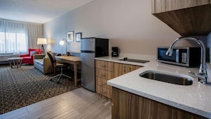A compact hotel room with a kitchenette, a desk, a chair, a sofa, and a window with curtains. - Days Inn & Suites by Wyndham Rochester Hills MI (Rochester)