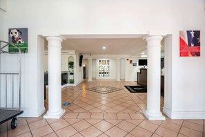 A hotel lobby with a tiled floor, white columns, a reception desk, and a view into the dining area.