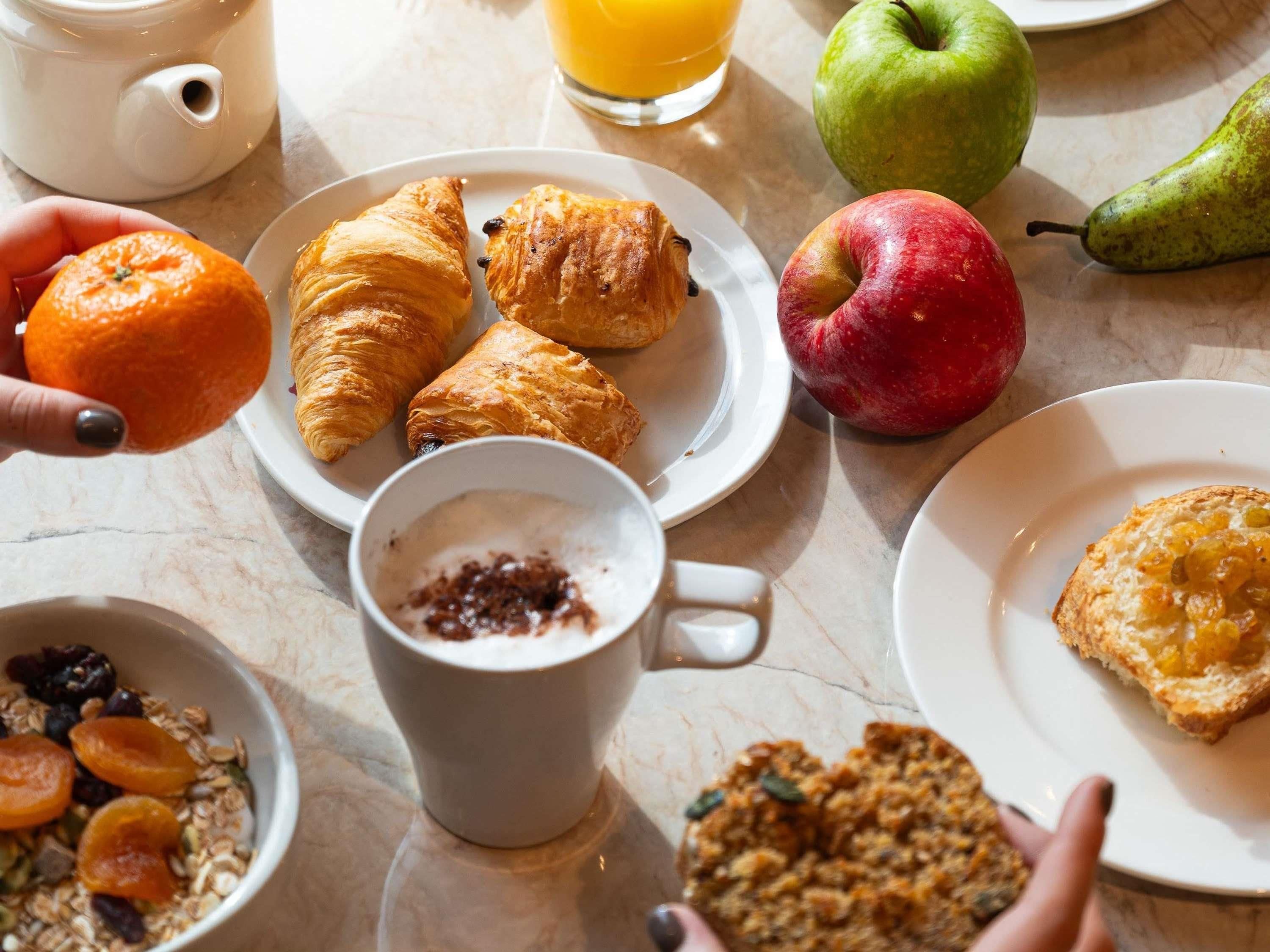 A breakfast setting with croissants, an apple, a pear, a glass of orange juice, a cup of coffee, and a bowl of granola.