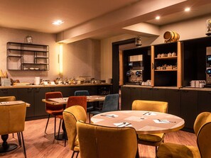 Un restaurante moderno con una zona central de comedor que cuenta con mesas redondas y sillas coloridas. Se ve un área de cocina con una máquina de café y estantes llenos de utensilios.