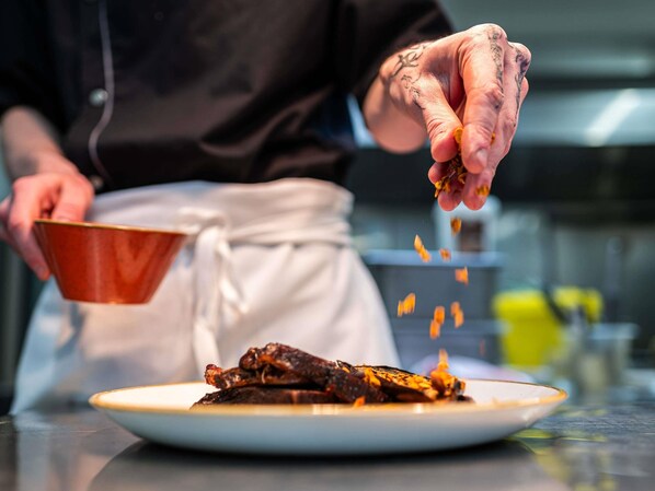 A person in a white apron holding a bowl, sprinkling seasoning on food. - Mercure Carcassonne la Cite Hotel (Carcassonne)