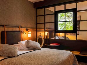 Habitación de hotel con cama, mesita de noche, ventana con vista a árboles y lámpara de pared.