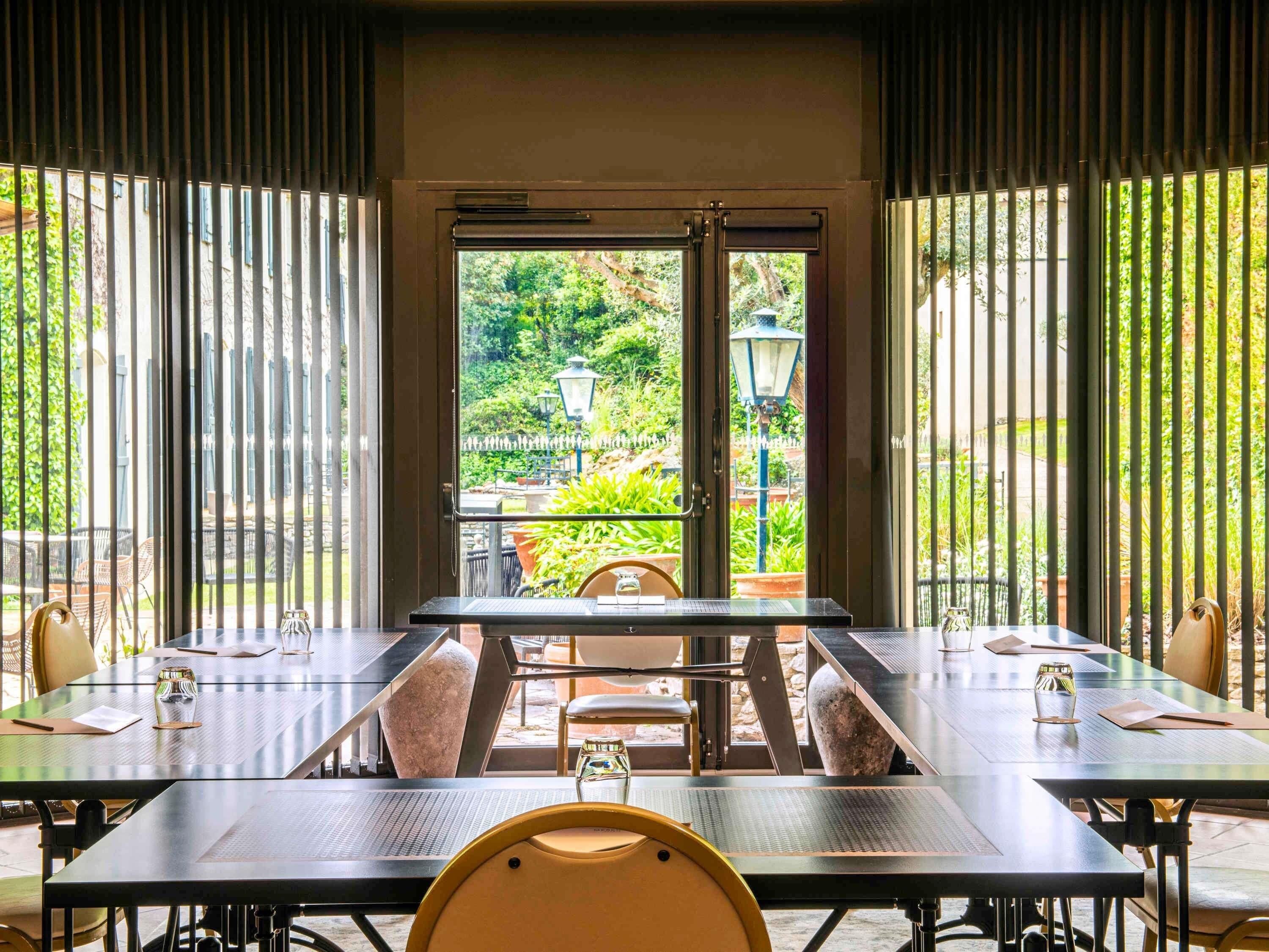 An outdoor dining area with tables and chairs, a view of greenery through a glass door, and a stone pillar.