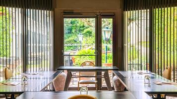 An outdoor dining area with tables and chairs, a view of greenery through a glass door, and a stone pillar.