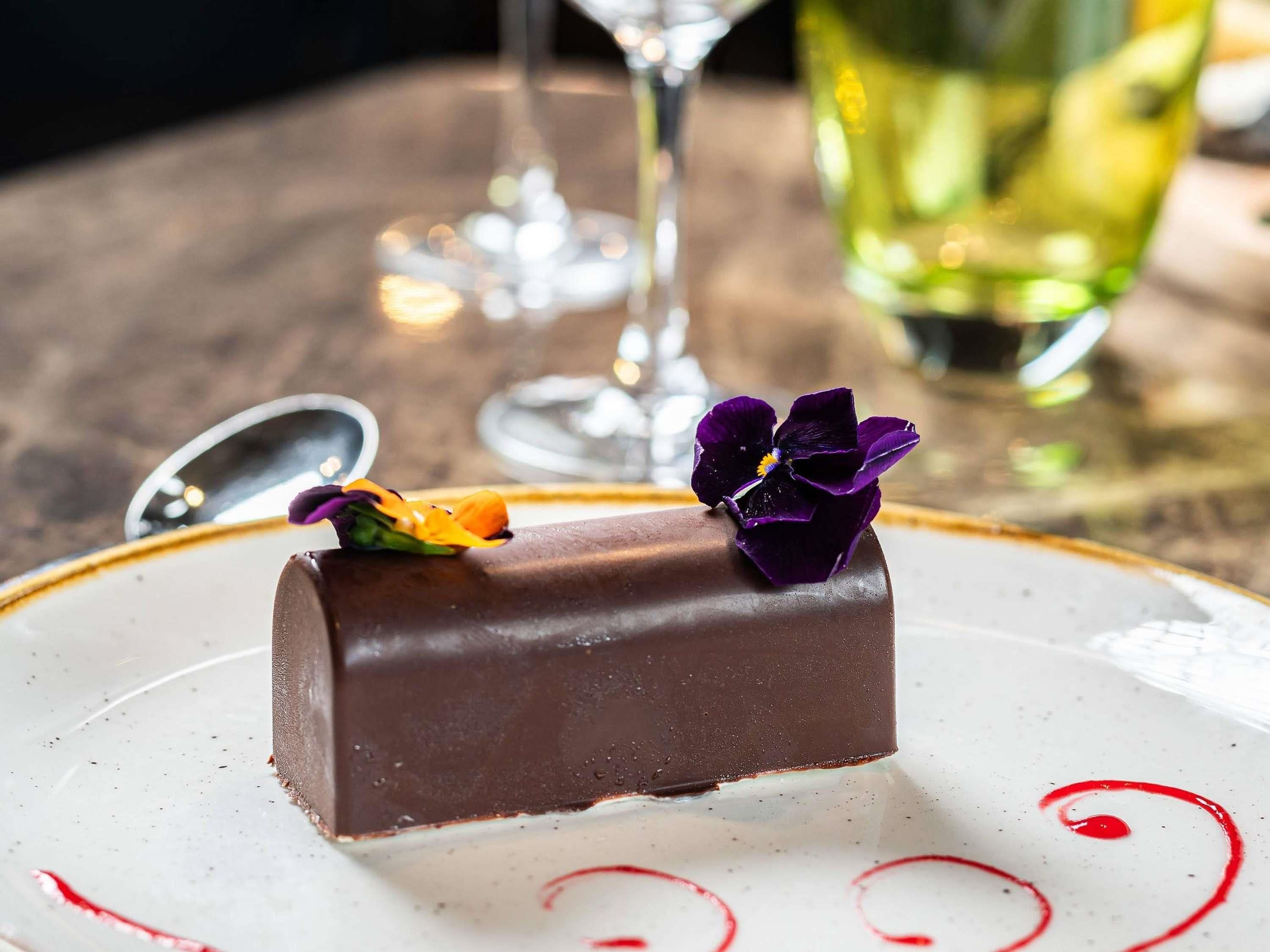 A chocolate dessert on a plate with a decorative flower and a glass of wine in the background.