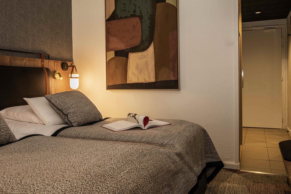 Une chambre à coucher comprenant un lit, un tableau abstrait accroché au mur, une lampe de chevet et une porte donnant sur une autre pièce.