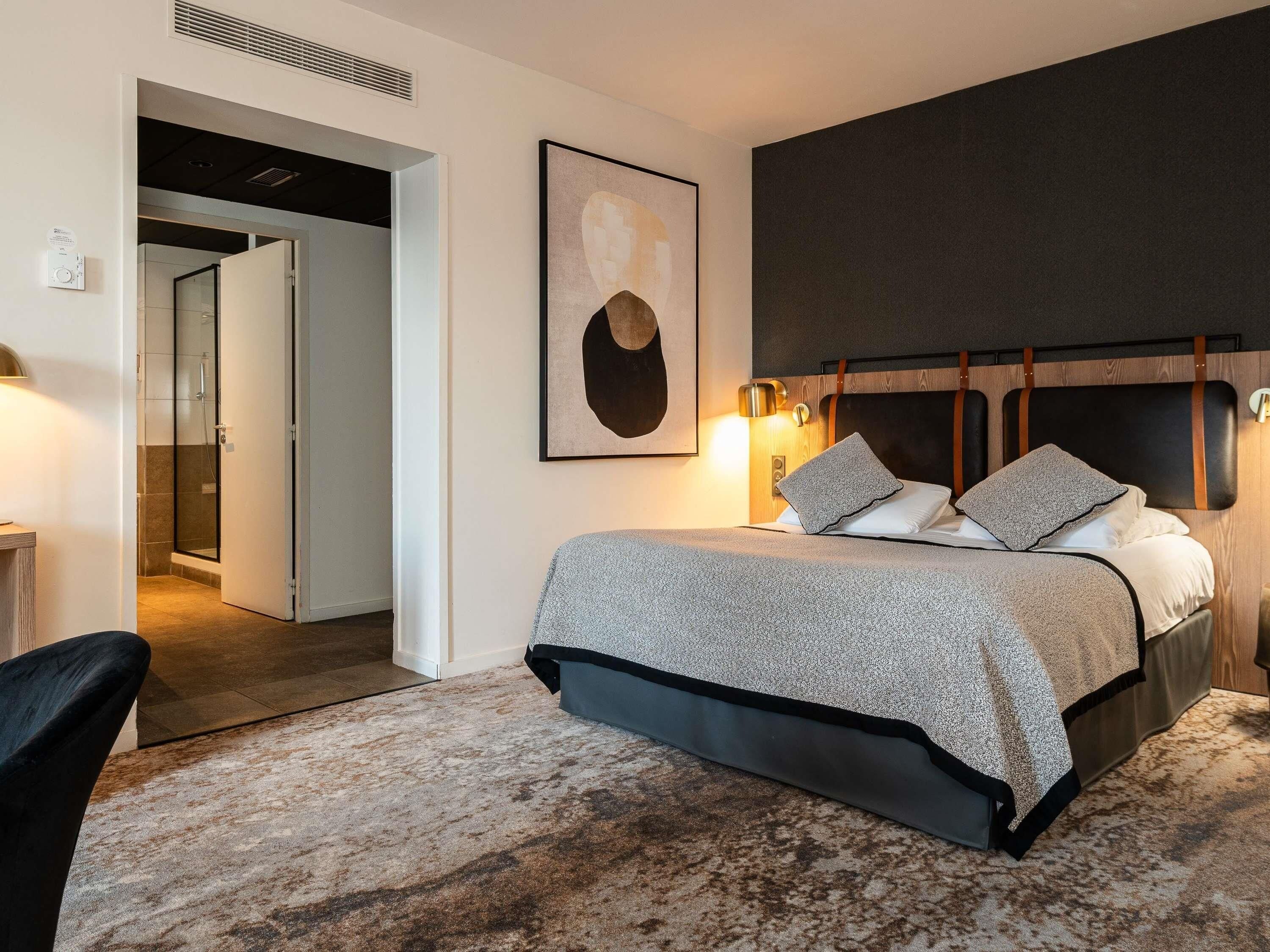 A modern bedroom with a large bed, a headboard with leather straps, a framed abstract painting, and a bedside lamp.