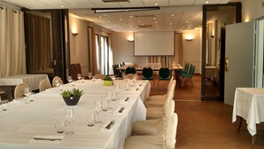 Un salón de conferencias con mesas largas dispuestas para una reunión, equipado con sillas, mesas y una pantalla para proyector.