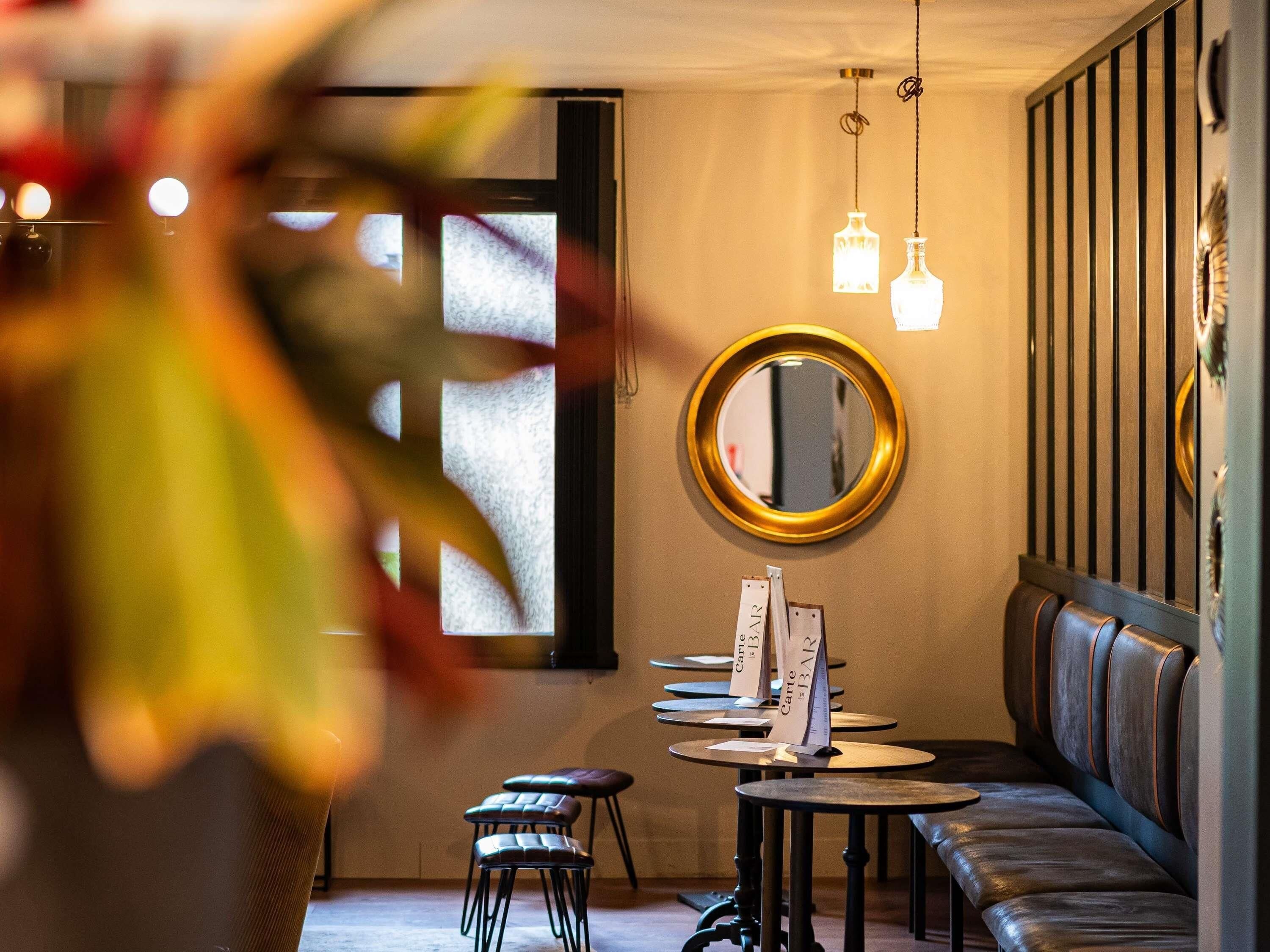 A restaurant interior with a long bench seating area, round tables, hanging pendant lights, and a wall-mounted mirror.