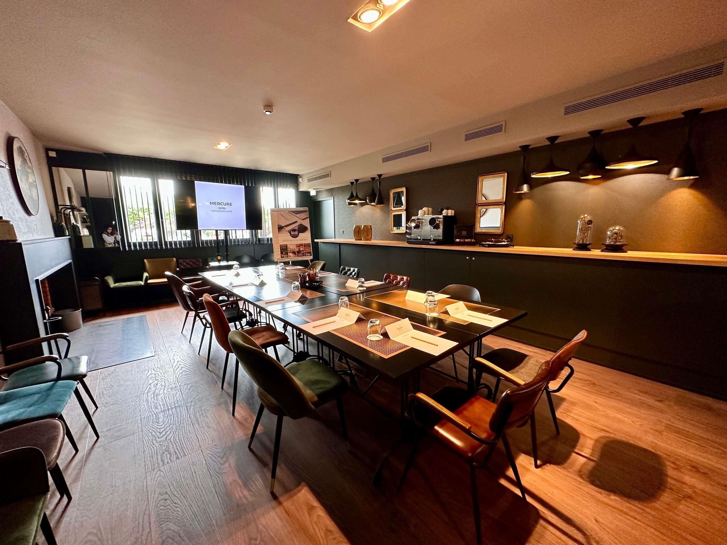 Uma sala de conferências com uma mesa comprida, cadeiras, uma tela de projetor e uma máquina de café.