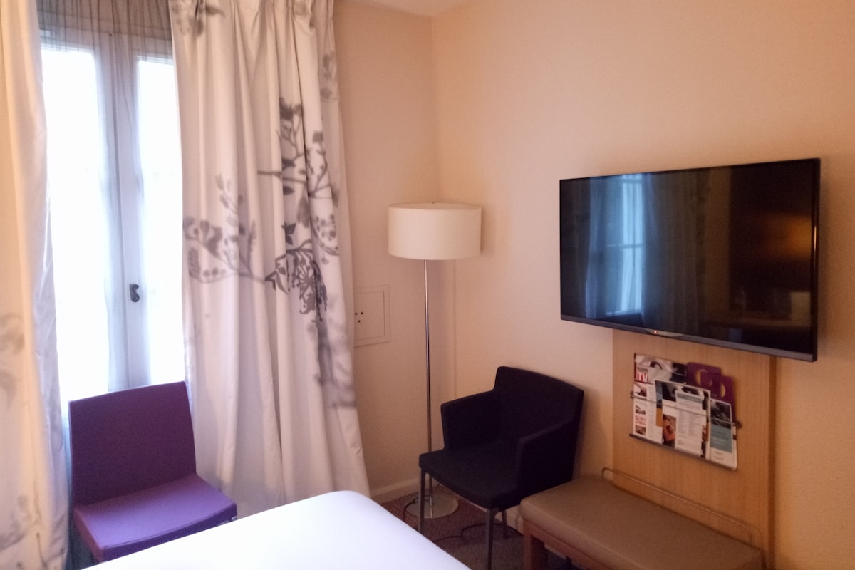 Quarto de hotel com cama, uma cadeira, televisão fixada na parede e janela com cortinas.