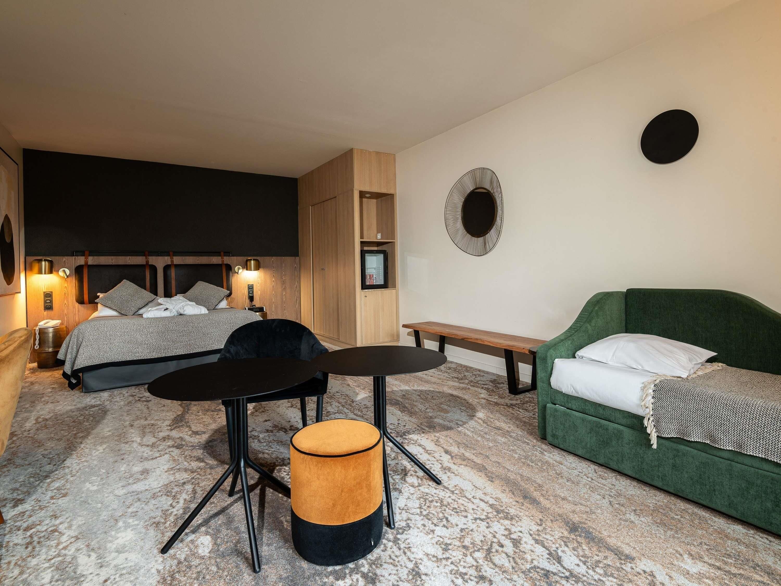 A hotel room with a bed, a green sofa, a round ottoman, a small table, and a bench.