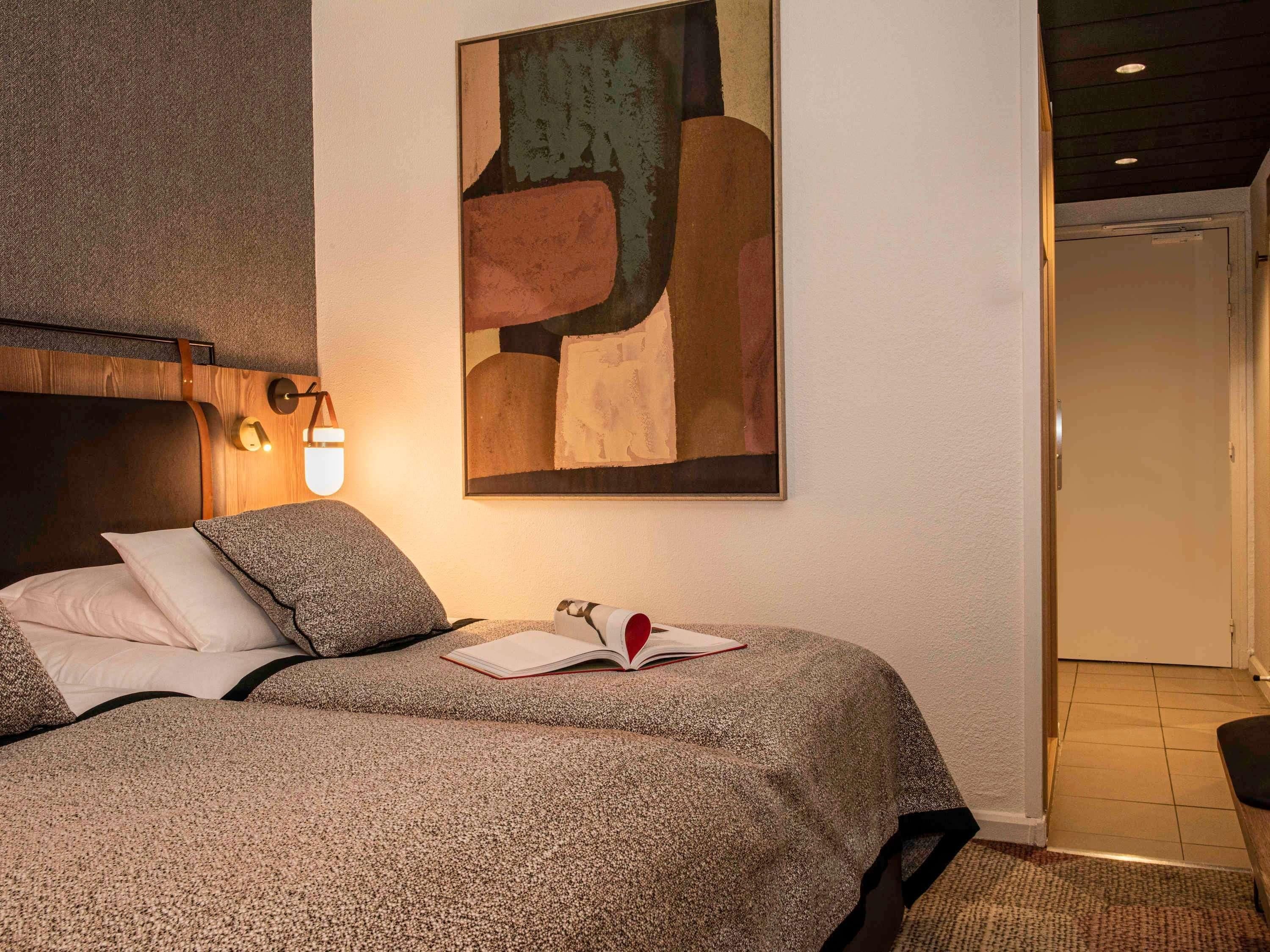 A bedroom with a bed, a wall-mounted abstract painting, a bedside table with a book, and a nightstand.