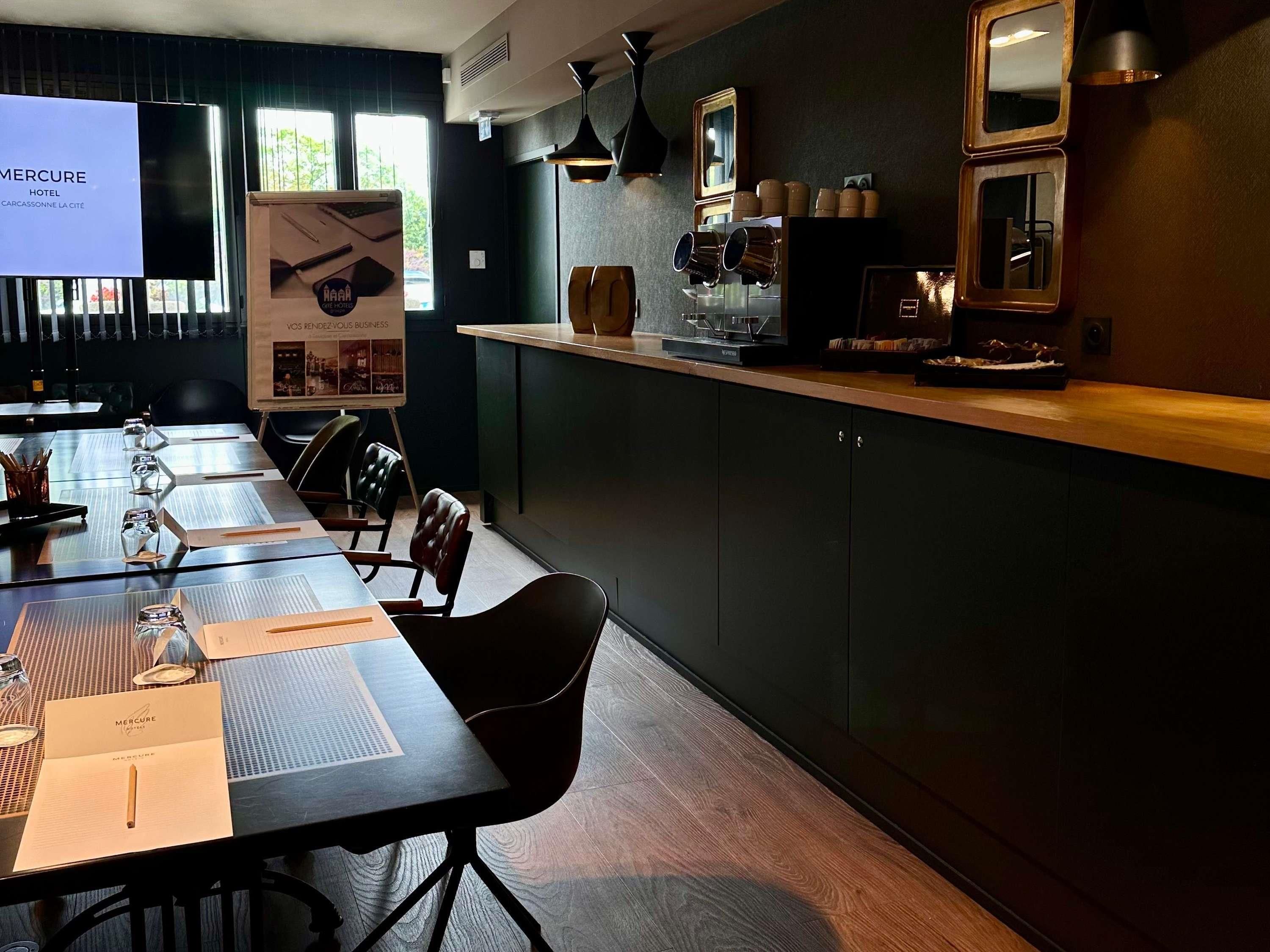 A conference room with a long table, chairs, a projector screen, and a wooden counter with kitchen appliances.