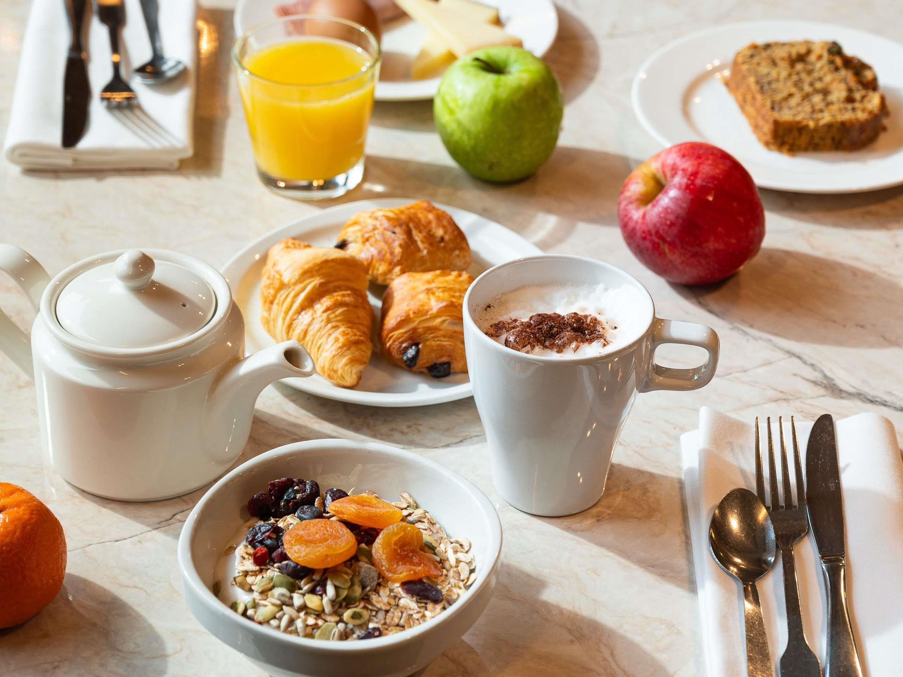 A breakfast setting with croissants, a bowl of granola, a mug of coffee, a glass of orange juice, and a plate with bread and an apple.