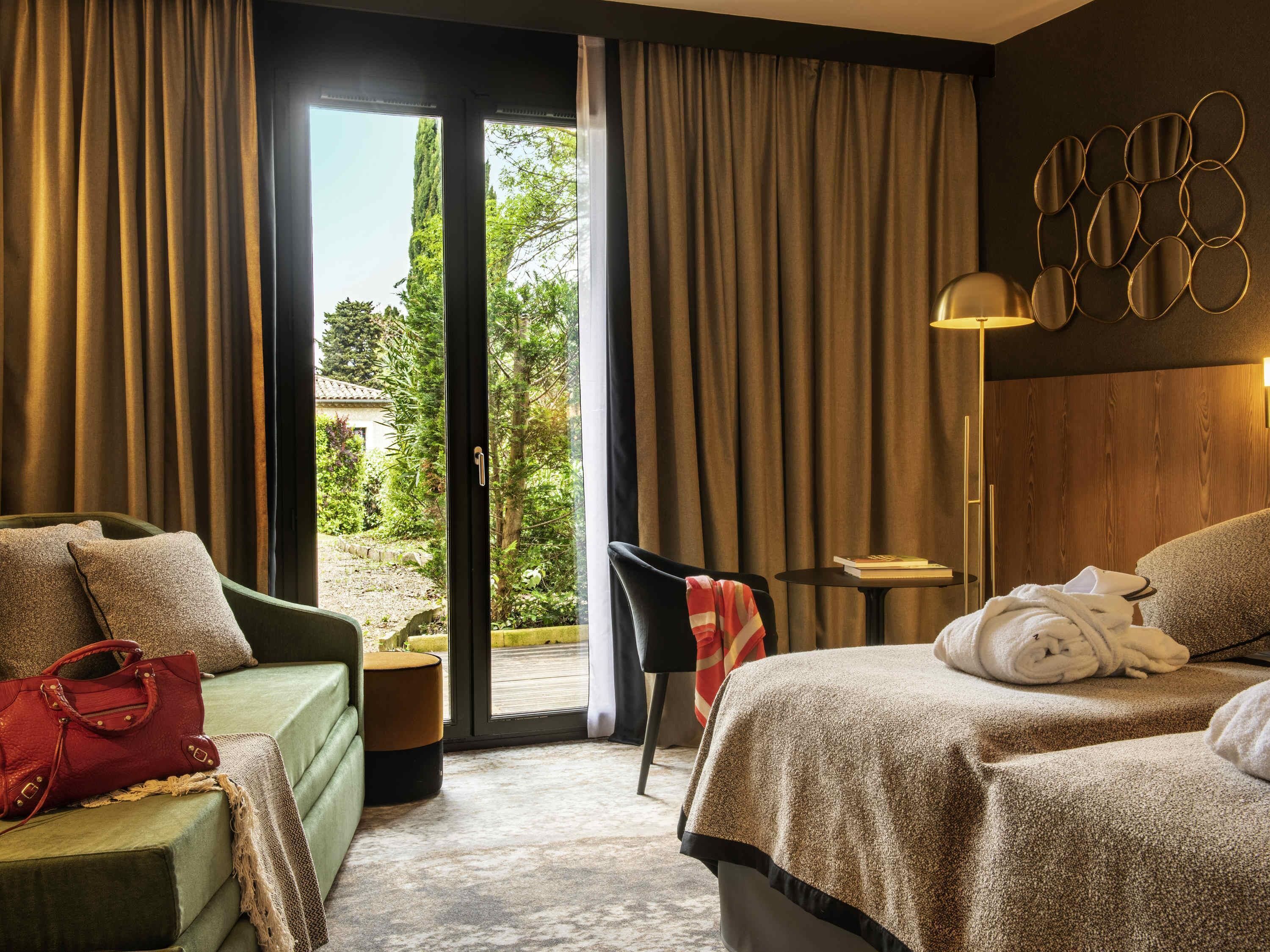 Habitación de hotel con cama, sofá verde, sillón, mesita y vistas a zonas verdes.