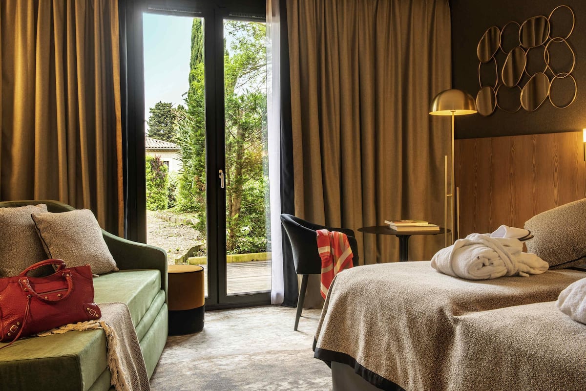 Habitación de hotel con cama, sofá verde, sillón, mesita y vistas a zonas verdes.
