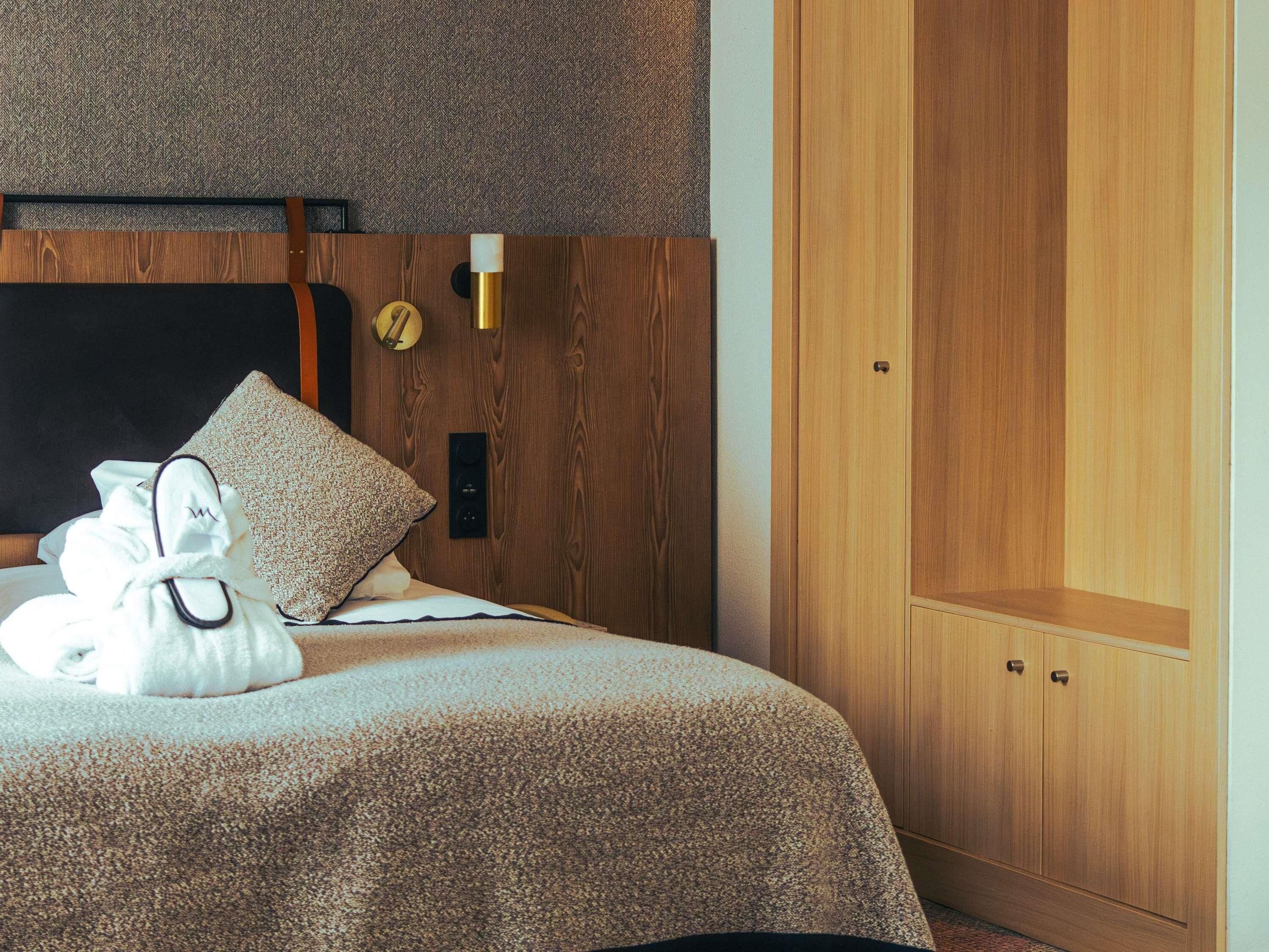 A hotel room with a bed, a wooden wardrobe, a wall clock, and a bedside table.