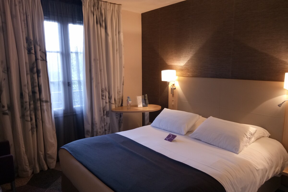 Quarto de hotel com cama, cortinas, mesa de cabeceira com abajur e luz na parede.