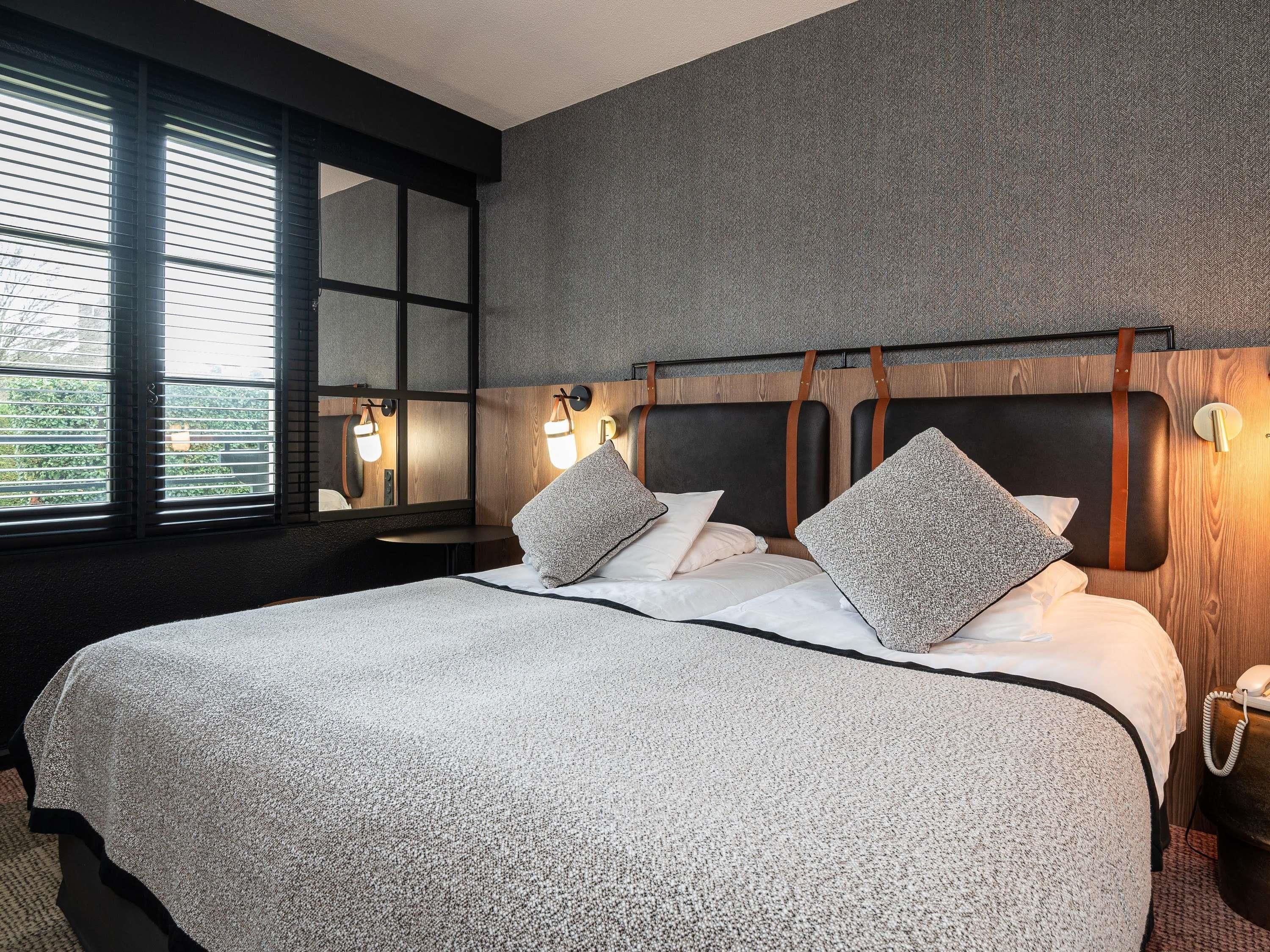 A hotel room with a large bed, two bedside lamps, a wooden headboard, and a window with blinds.