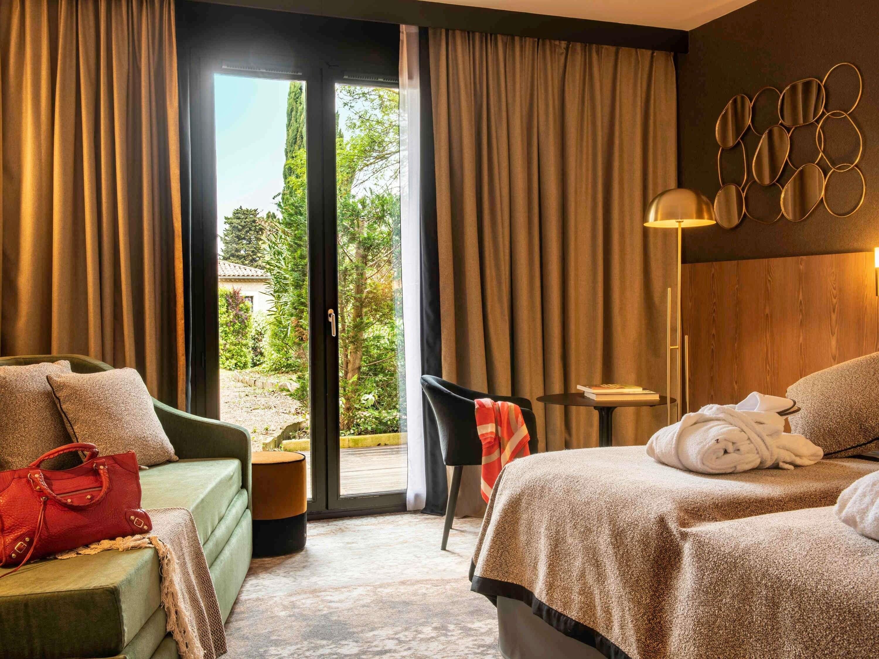 Habitación de hotel con una cama, un sofá verde, una mesita y una silla. Se ve un jardín a través de la ventana.