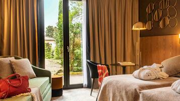 A hotel room with a bed, a green sofa, a small table, and a chair. There is a view of a garden through the window.