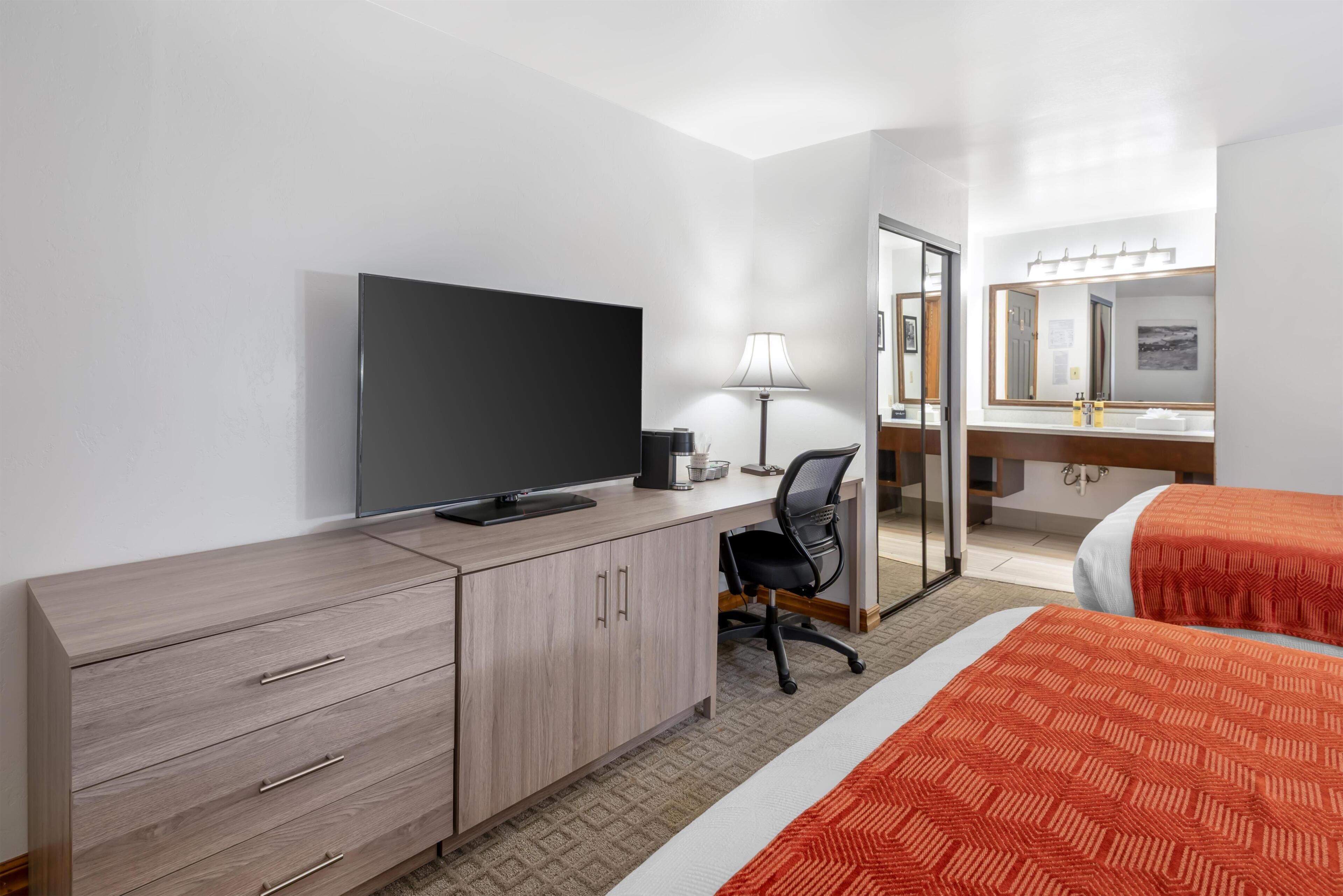 Ein Hotelzimmer mit Fernseher, Schreibtisch, Stuhl, zwei Betten und einem Badezimmer-Spiegel.