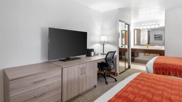 A hotel room with a TV, desk, chair, two beds, and a bathroom mirror.