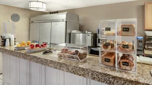 A hotel kitchen with a granite countertop, a stainless steel refrigerator, a clock, and a bread display.