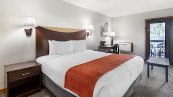 A hotel room with a bed, bedside table, lamp, desk, chair, and a view of the outdoors.