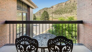 A balcony with a table and chairs, a view of a hilly landscape, and a brick building.