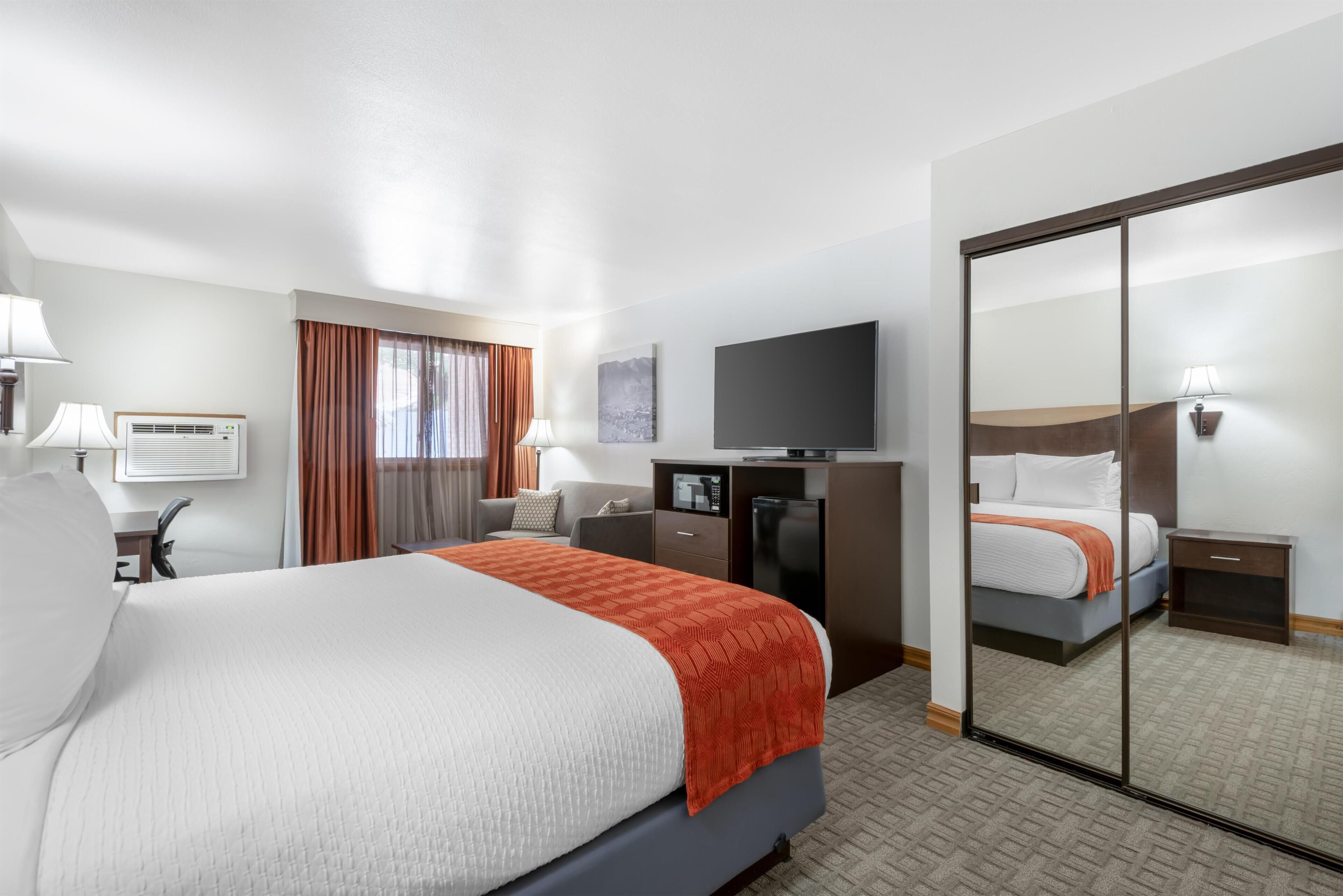A hotel room with a large bed, a desk with a computer, a TV, a mirror, and a wardrobe.