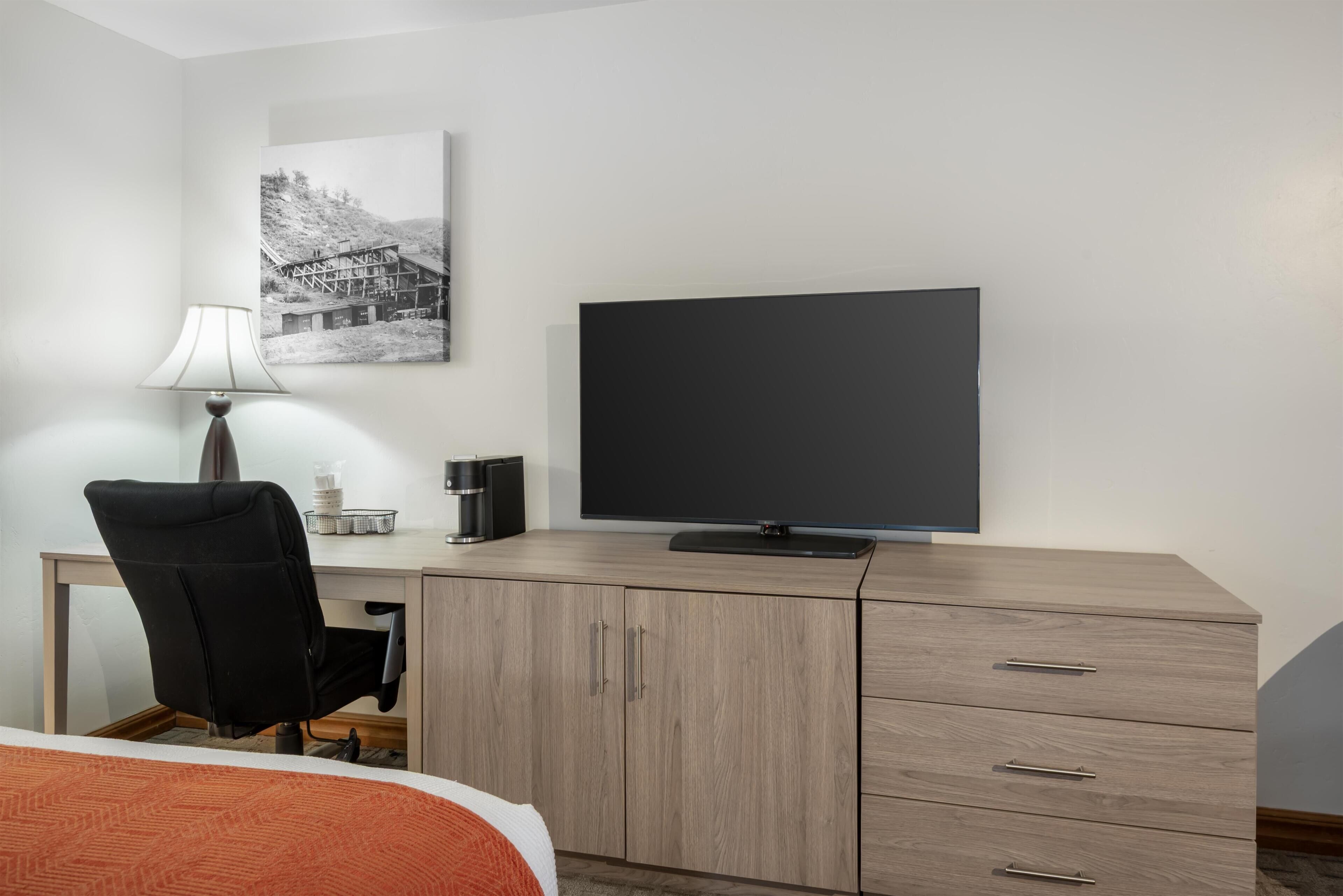 A hotel room with a bed, a desk with a chair, a television, a lamp, and a picture on the wall.
