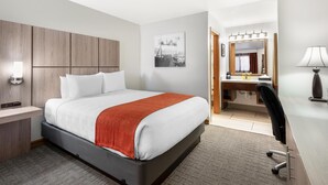 A hotel room with a bed, bedside table, lamp, desk, chair, and a bathroom with a mirror and sink.